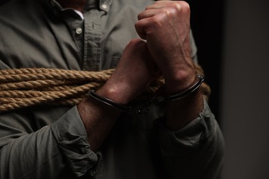 Photo of Handcuffed victim tied with rope on dark background, closeup. Hostage taking