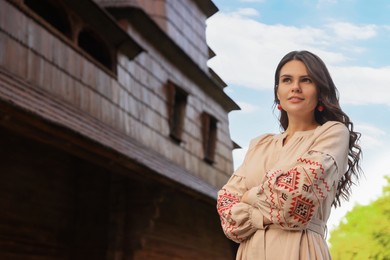 Beautiful woman wearing embroidered dress near old wooden church in village. Ukrainian national clothes