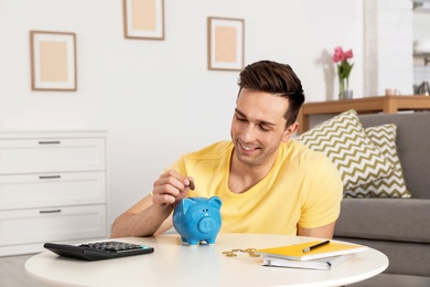 Happy man putting coin into piggy bank at table in living room. Saving money