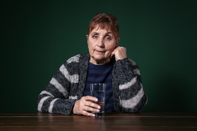 Poor senior woman with glass of water sitting at table against color background