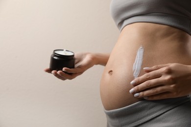 Pregnant woman applying cosmetic product on belly against beige background, closeup