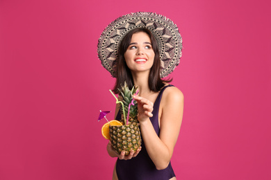 Beautiful woman in stylish swimsuit holding tropical cocktail on pink background