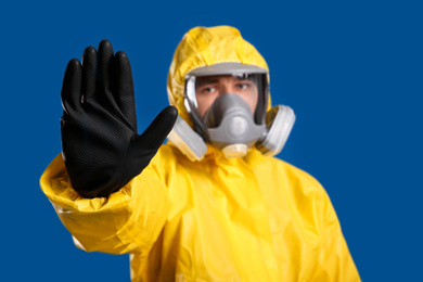 Man in chemical protective suit making stop gesture against blue background, focus on hand. Virus research