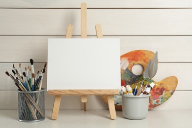 Easel with blank canvas, brushes, paints and palette on white wooden table