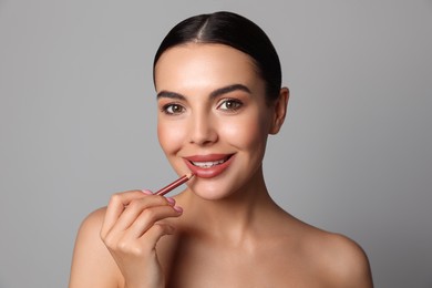 Pretty young woman applying beautiful nude lip pencil on grey background