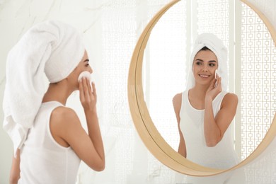 Happy young woman cleaning face with cotton pad near mirror in bathroom