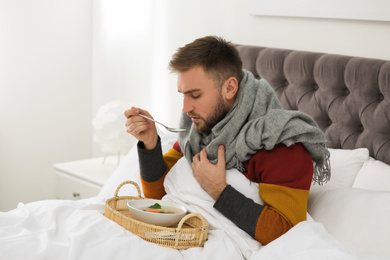 Sick young man with bowl of tasty soup in bed at home