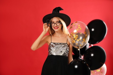 Beautiful woman in witch costume with balloons on red background, space for text. Halloween party