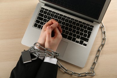 Man showing chained hands near laptop at wooden table, top view. Internet addiction