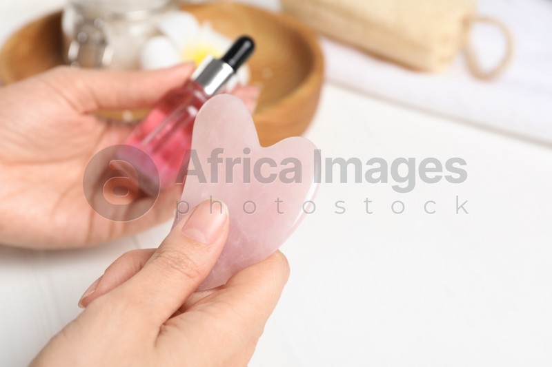 Photo of Woman holding gua sha tool and cosmetic at white wooden table, closeup. Space for text