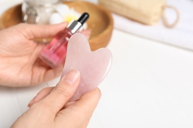 Photo of Woman holding gua sha tool and cosmetic at white wooden table, closeup. Space for text