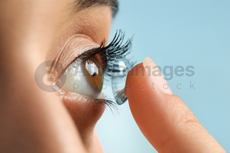 Young woman putting contact lens in her eye on light blue background, closeup