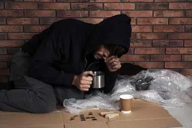 Poor man with piece of bread and mug near brick wall