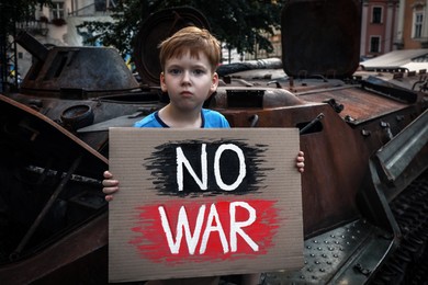 Sad boy holding poster No War near broken military tank outdoors