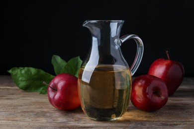 Jug of tasty juice and fresh red apples on wooden table against black background