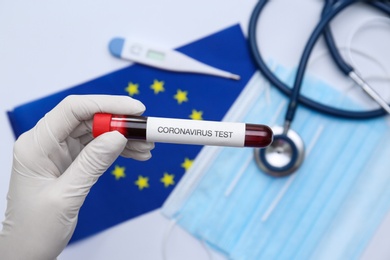 Doctor holding sample tube with label Coronavirus Test above medical items and European Union flag, closeup