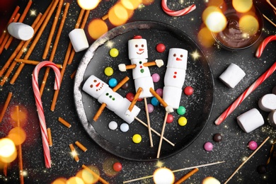 Flat lay composition with funny snowmen made of marshmallows on black table. Bokeh effect