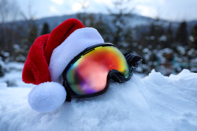 Stylish ski goggles with Santa hat on snow outdoors. Winter sport equipment