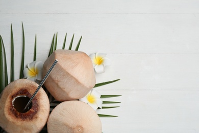 Young peeled coconuts with straw, tropical leaf and flowers on white wooden table, flat lay. Space for text