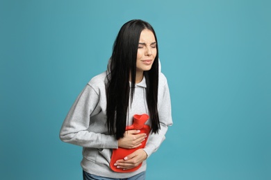 Woman using hot water bottle to relieve abdominal pain on light blue background