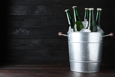 Photo of Metal bucket with bottles of beer and ice cubes on wooden table against dark background, space for text