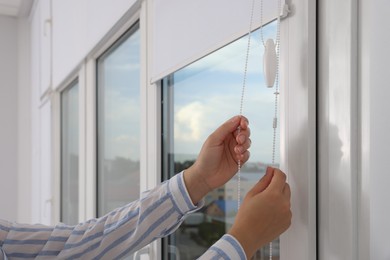 Photo of Woman opening white roller blinds on window indoors, closeup