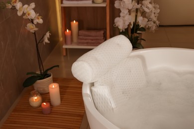 Tub with foamy water and soft bath pillow surrounded by candles indoors
