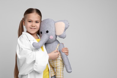 Photo of Little girl in medical uniform with toy elephant on light grey background. Space for text