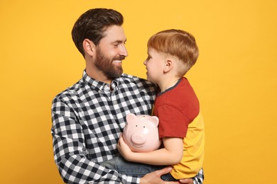 Father and his son with ceramic piggy bank on orange background