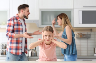 Little unhappy girl sitting at table while parents arguing on kitchen