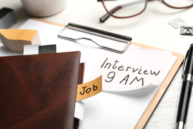 Photo of Clipboard with reminder note about job interview and stationery on table, closeup