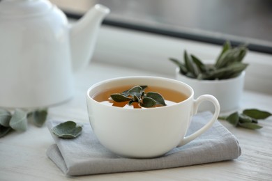 Cup of sage tea and green leaves on white wooden windowsill