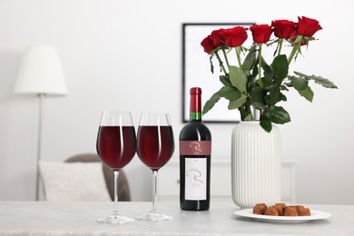 Bottle, glasses of red wine, chocolate truffles and vase with roses on table in room. Romantic date