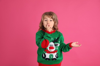 Photo of Cute little girl in green Christmas sweater showing silence gesture against pink background