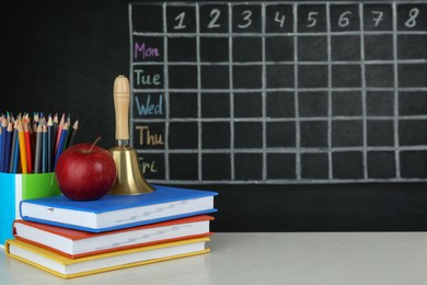 Apple, bell and different stationery on white wooden table near blackboard with chalked school timetable