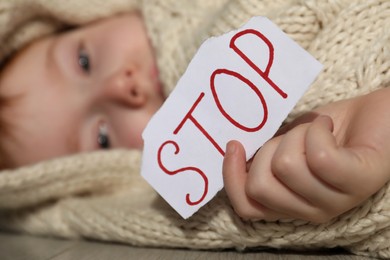 Photo of Scared little boy wrapped in blanket holding piece of paper with word Stop on floor, closeup. Domestic violence concept