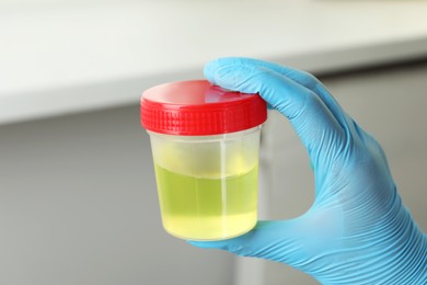 Doctor wearing glove holding container with urine sample for analysis indoors, closeup