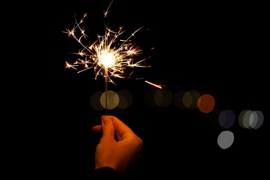 Woman holding bright burning sparklers against blurred lights, closeup. Space for text