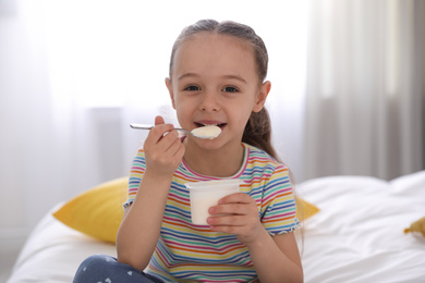 Cute little girl eating tasty yogurt on bed at home