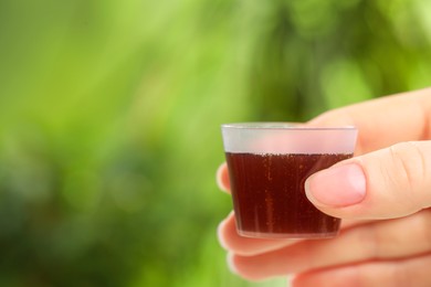 Photo of Woman holding measuring cup with syrup on blurred background, closeup and space for text. Cold medicine