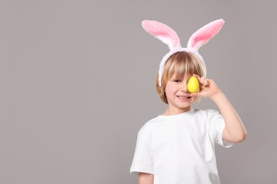 Photo of Happy boy in bunny ears headband holding painted Easter egg on grey background. Space for text