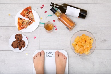 Woman weighing on scales surrounded by junk food and alcohol after party indoors, top view