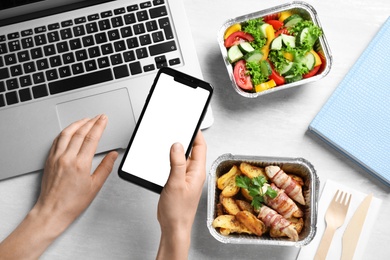 Top view of woman holding smartphone over white wooden table with lunchboxes,  mockup for design. Healthy food delivery