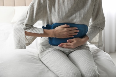 Woman using hot water bottle to relieve abdominal pain on bed at home, closeup