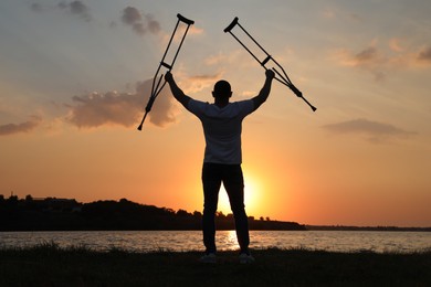 Man raising hands with underarm crutches up to sky near river at sunset, back view. Healing miracle