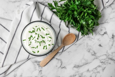 Glass bowl of fresh sour cream with parsley and wooden spoon on marble table, flat lay. Space for text