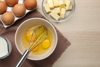 Whisk and eggs in bowl, butter, milk on wooden table, flat lay. Space for text