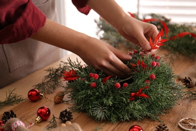 Florist making beautiful Christmas wreath with berries and red leaves at wooden table indoors, closeup