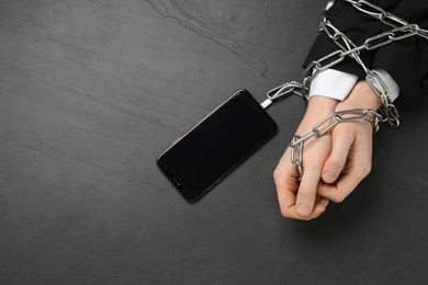 Man showing chained hands to smartphone at black table, top view. Internet addiction