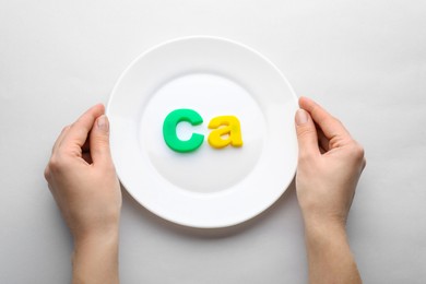 Woman holding plate with calcium symbol made of colorful letters on light grey background, top view